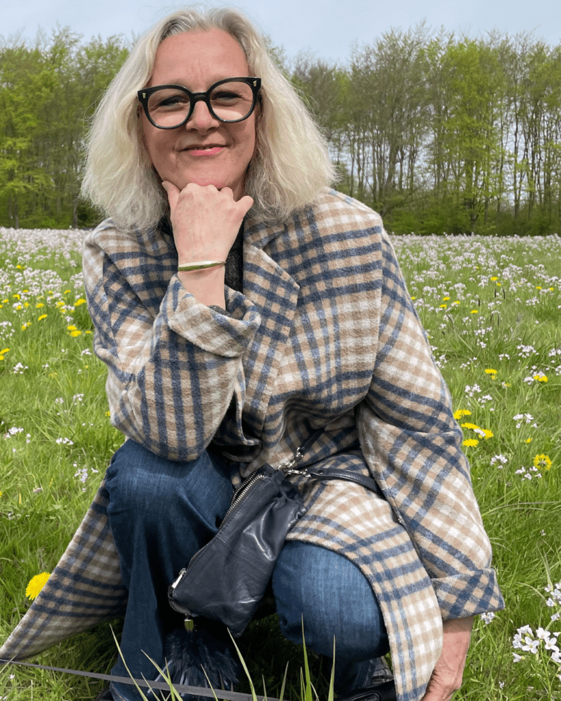 Birgitte-med-frakke-paa-en-mark-med-blomster-Blog-Vi-arbejder-i-fire-lag-af-bevidsthed.